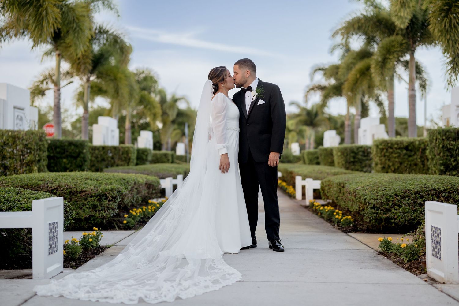 A couple shares a romantic moment on a palm-lined path during their elegant outdoor wedding ceremony.