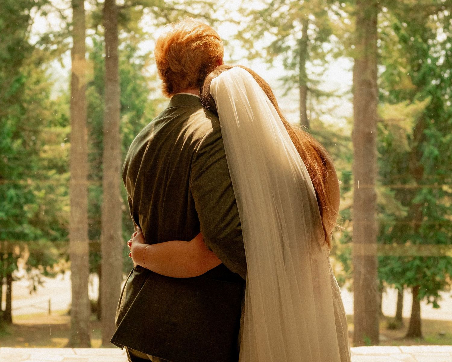 A couple embraces in a sun-dappled forest setting with a flowing white veil.