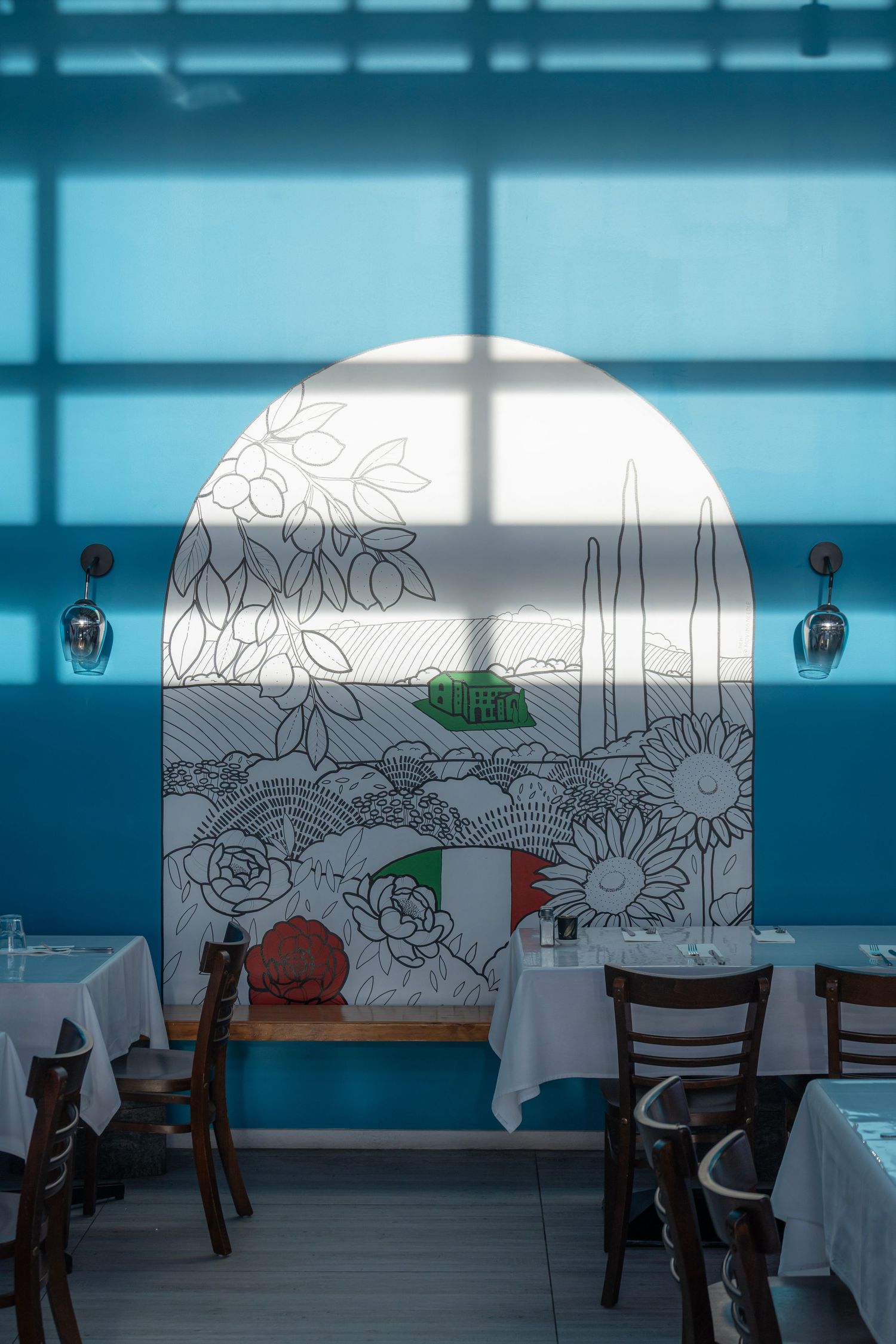 Blue restaurant wall with a painted Tuscan arch and mural casting a midday window shadow.