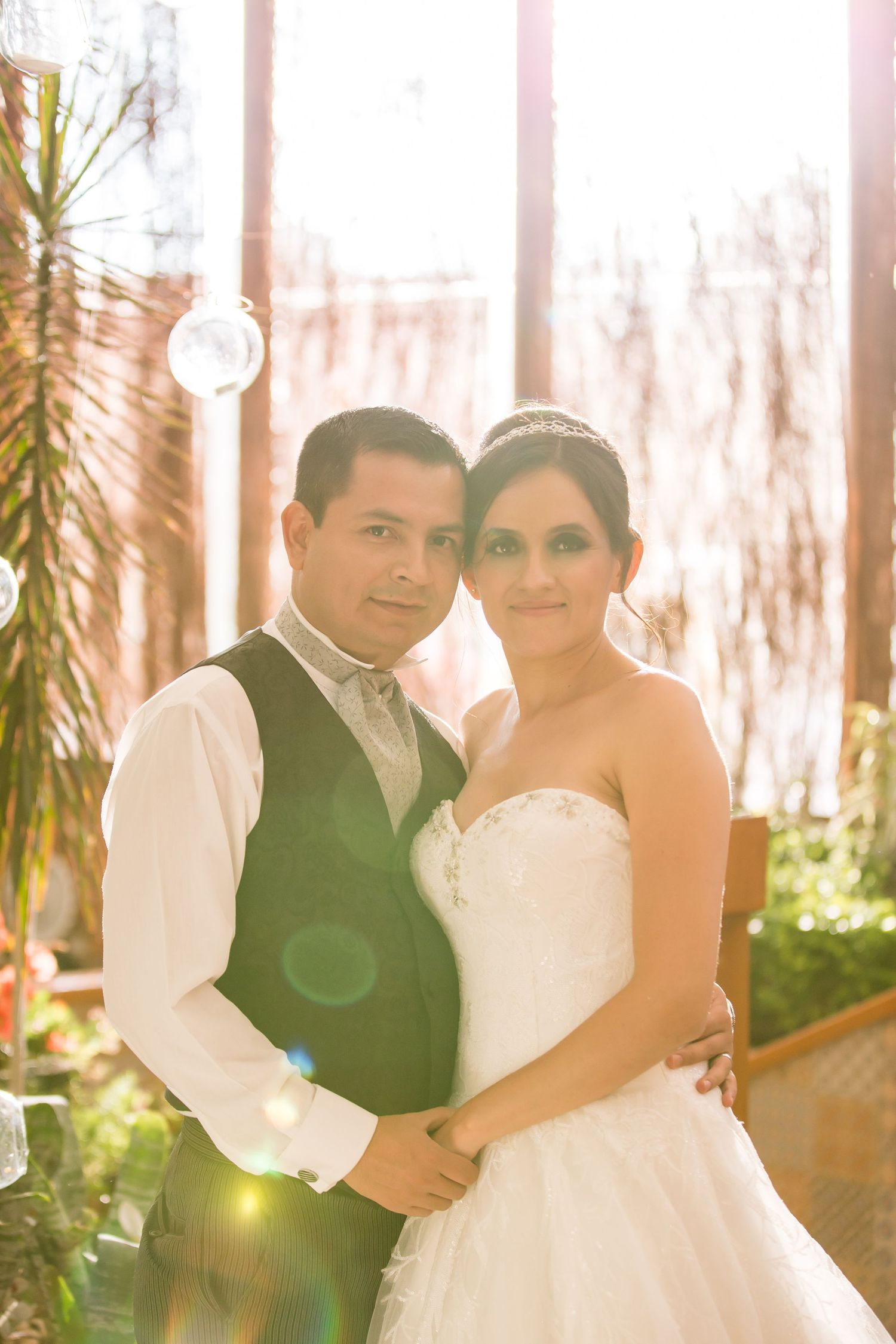 A couple poses in wedding attire with soft sunlit glow filtering through windows and palm trees in the background.