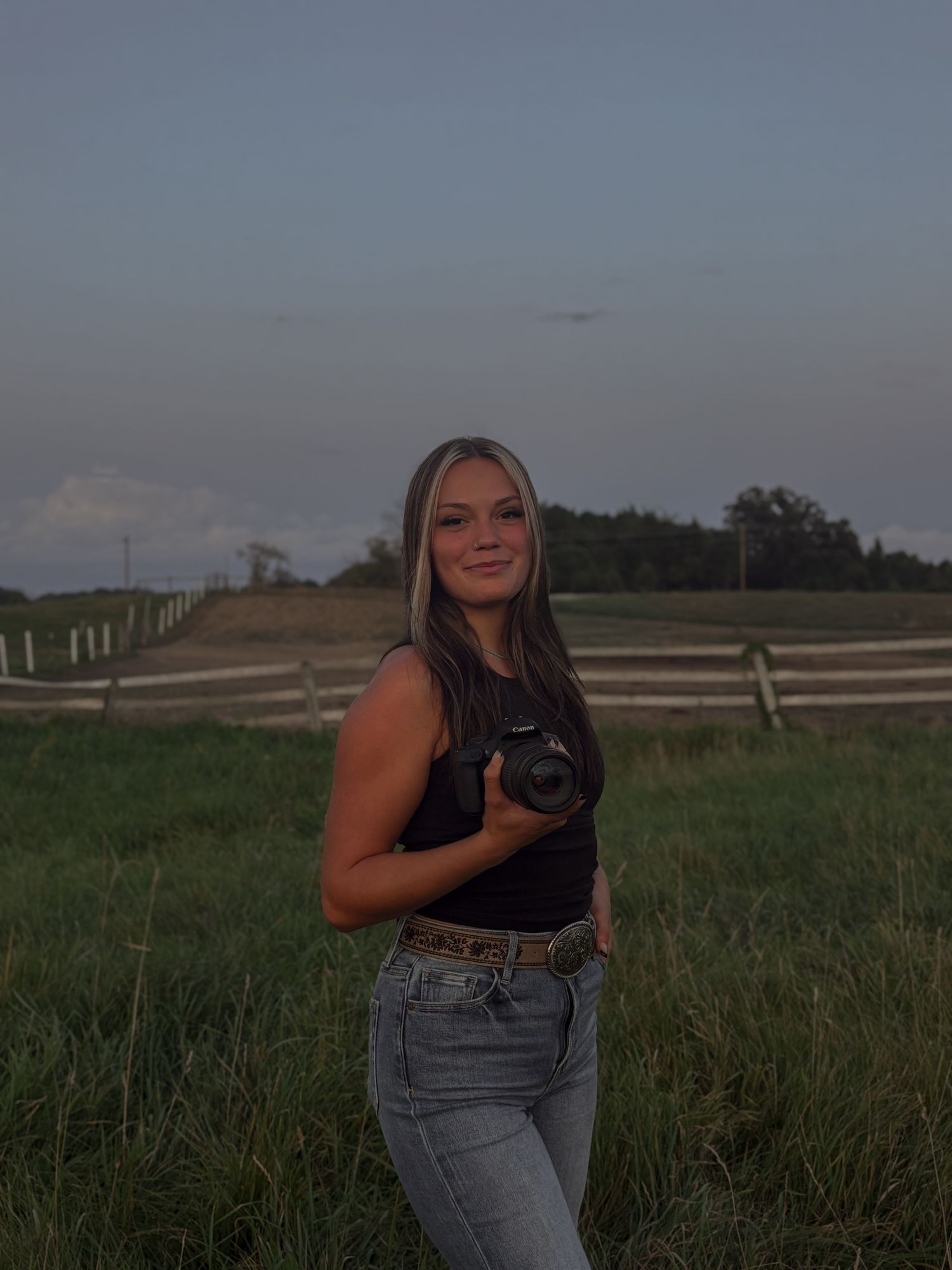 A photographer with camera stands in a field with wooden fences at dusk.