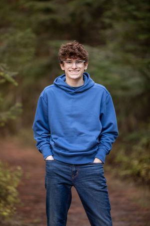 A person wearing a blue hoodie and jeans stands smiling on a forest path with hands in pockets.