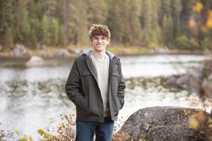A person in a black jacket stands on a rocky shore beside a serene lake with evergreen forest in the background.