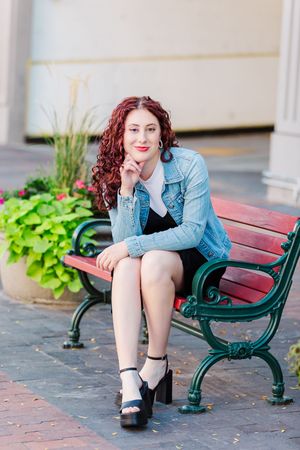 Person in denim jacket sitting on green park bench with decorative plants nearby.