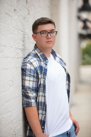 Young person in glasses wearing a blue plaid short-sleeve button up shirt over white t-shirt poses against textured wall.