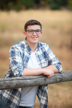 A young person in glasses and plaid shirt leans casually against a wooden fence in an outdoor setting.
