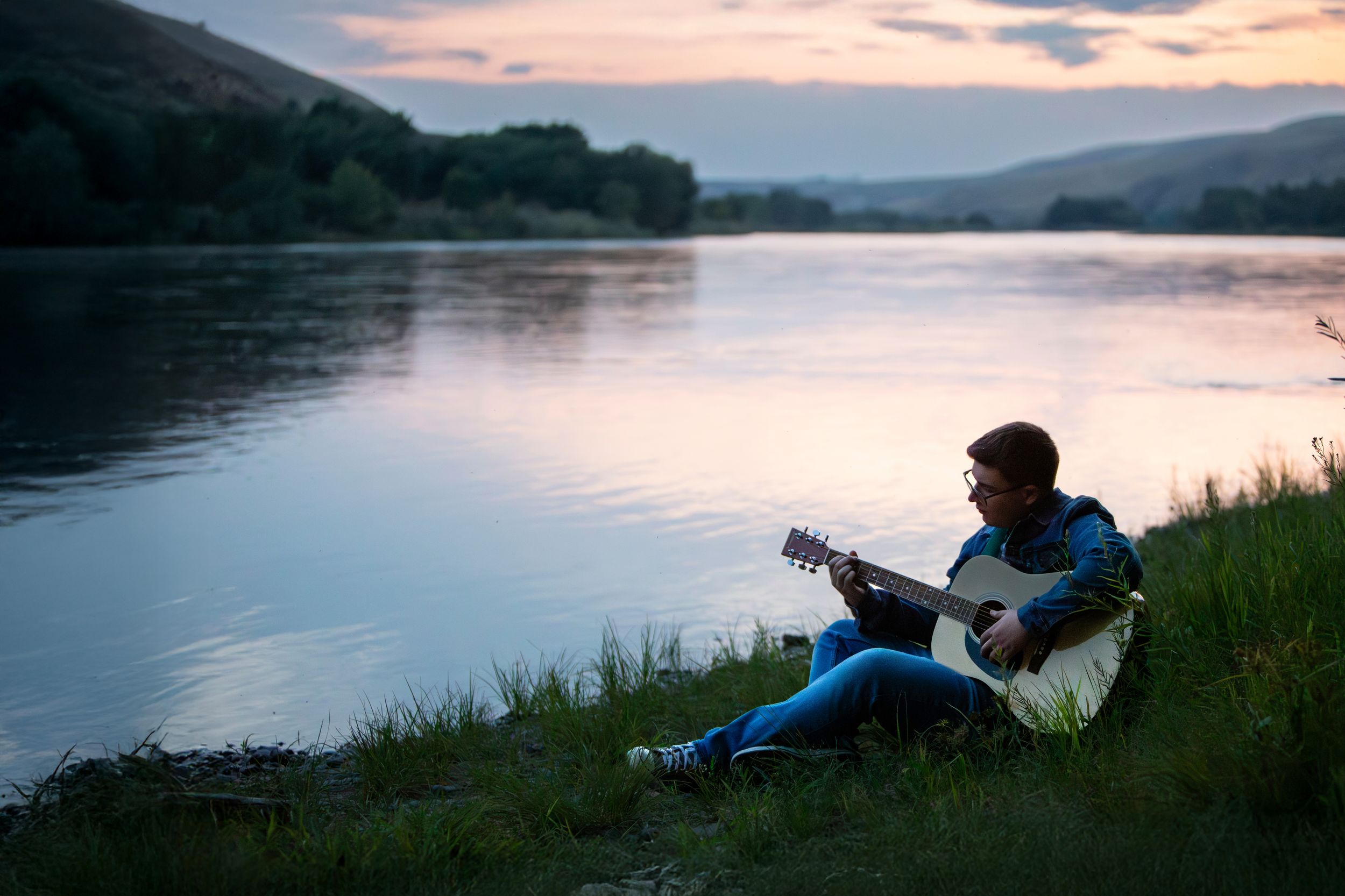 Silhouetted figure plays acoustic guitar by tranquil lake at sunset with mountains in background.