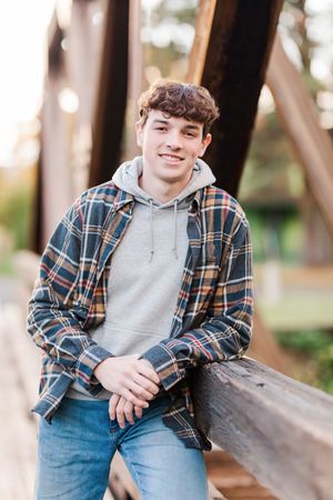 Person in plaid shirt and gray hoodie poses against wooden structure outdoors.