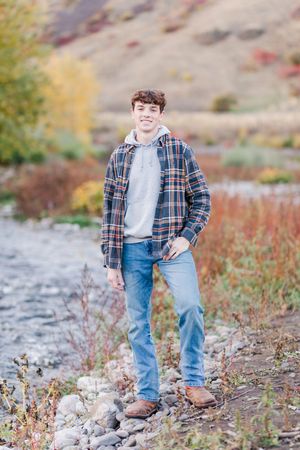 Person in plaid shirt and jeans poses on rocky terrain with autumn foliage in background.