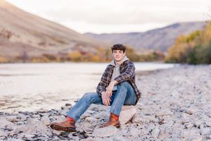 Person relaxing on rocky shore with mountains and lake in background.