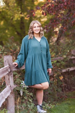 Person wearing a teal dress and sneakers posing by a rustic wooden fence in autumn.