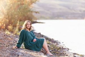 Person in a teal dress sitting by a rocky shoreline at sunset.