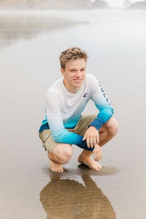 Person in blue ombre shirt crouching on wet sand at the beach.