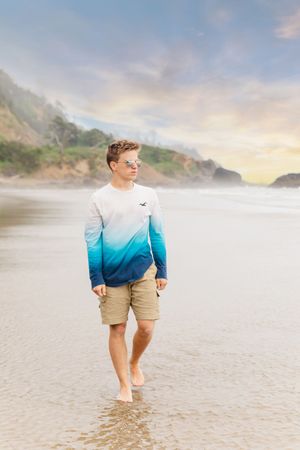 Person walking barefoot on beach wearing blue ombre shirt and khaki shorts at sunset.