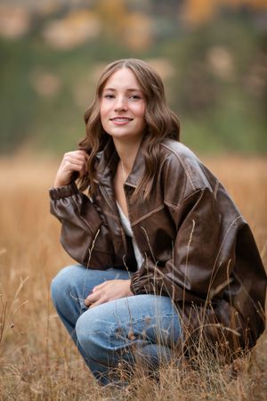 A person in a brown leather jacket sits in tall autumn grass during an outdoor portrait session.