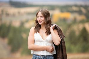 A woman in a white top and denim stands in a natural outdoor setting with golden sunlight and foliage in the background.