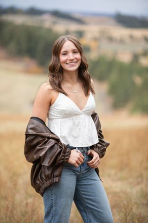 A woman in a white top and brown leather jacket poses in a sunny field with natural golden light.