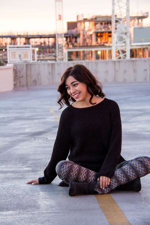 Person in black sweater and patterned pants sitting on rooftop at sunset.