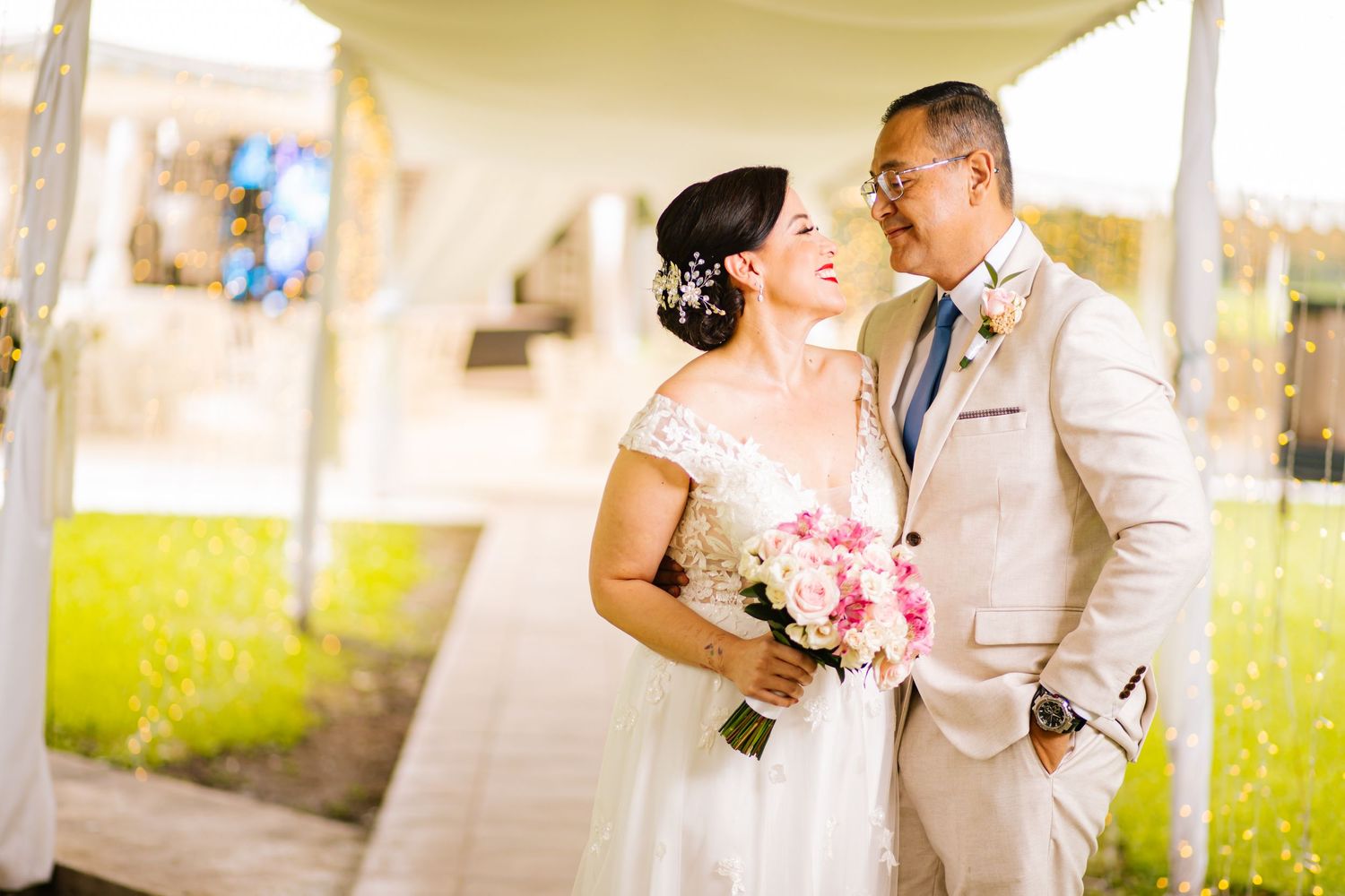 A newlywed couple shares a romantic moment under an outdoor archway, with the bride holding a pink bouquet.