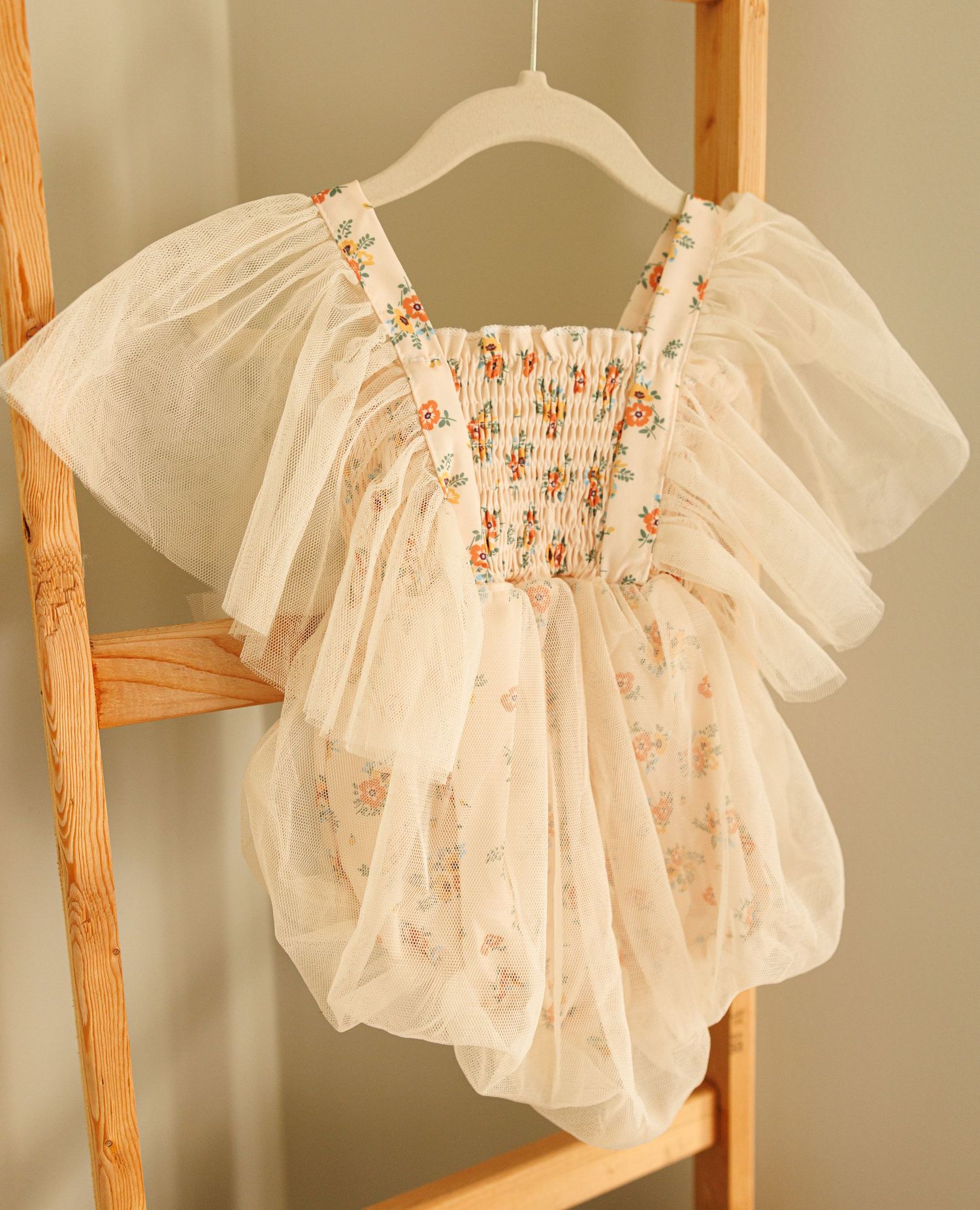 Delicate cream-colored tulle dress with flutter sleeves and smocked bodice featuring tiny floral embroidery hangs on white hanger.