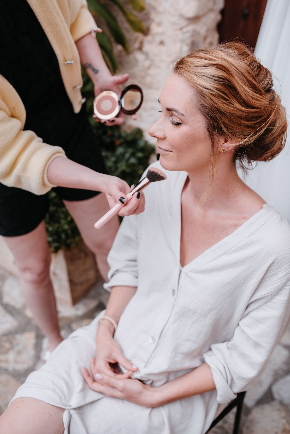 Hochzeitsvorbereitungen, die die Braut in einem weißen Kleid zeigen. La Chia trägt ihr Make-Up auf im Innenhof der Finca Xiclati, Mallorca.