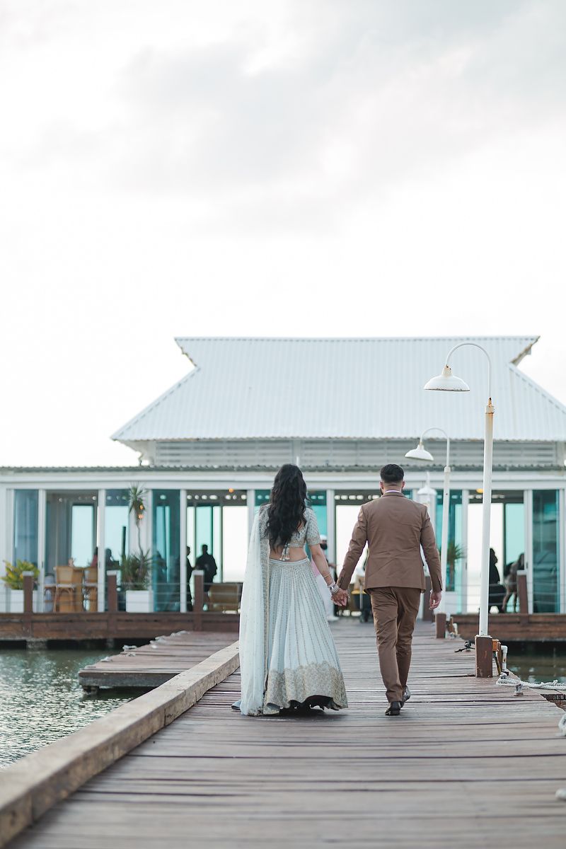Wedding couple walking on wooden pier towards white waterfront pavilion.