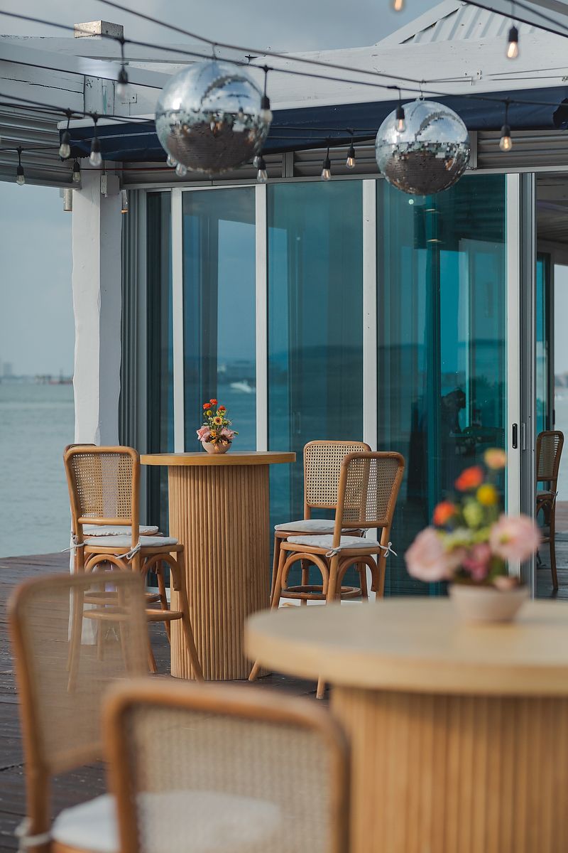 Modern coastal restaurant interior with wooden chairs and glass walls offering panoramic waterfront views.