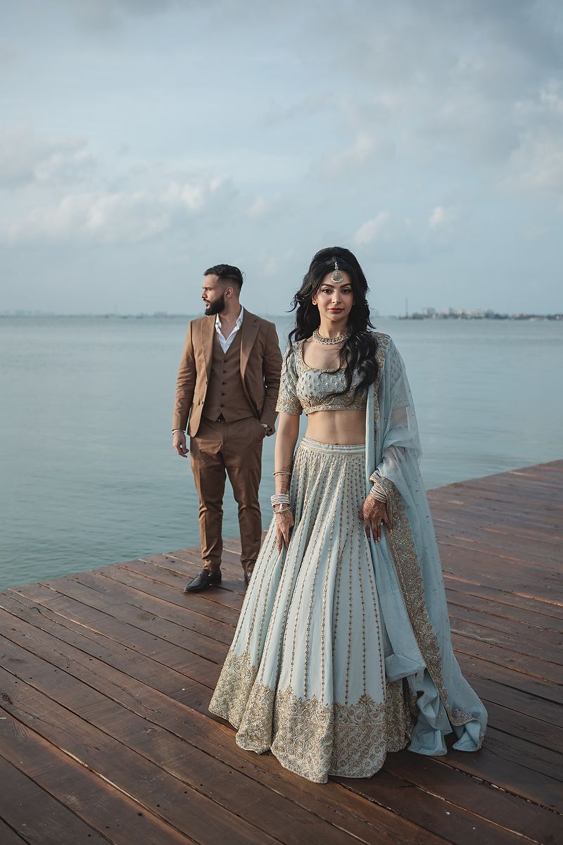 A couple in elegant Indian wedding attire posing on a waterfront wooden deck with a serene ocean backdrop.