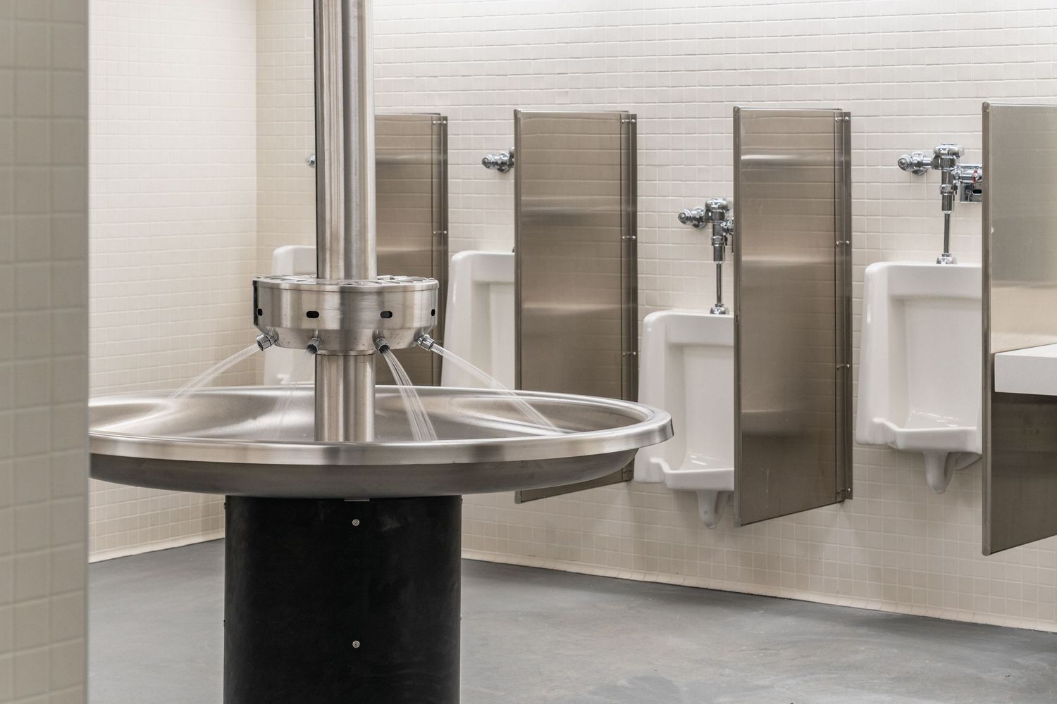 Circular wash basin with stainless steel fixtures and urinal dividers in a commercial restroom.