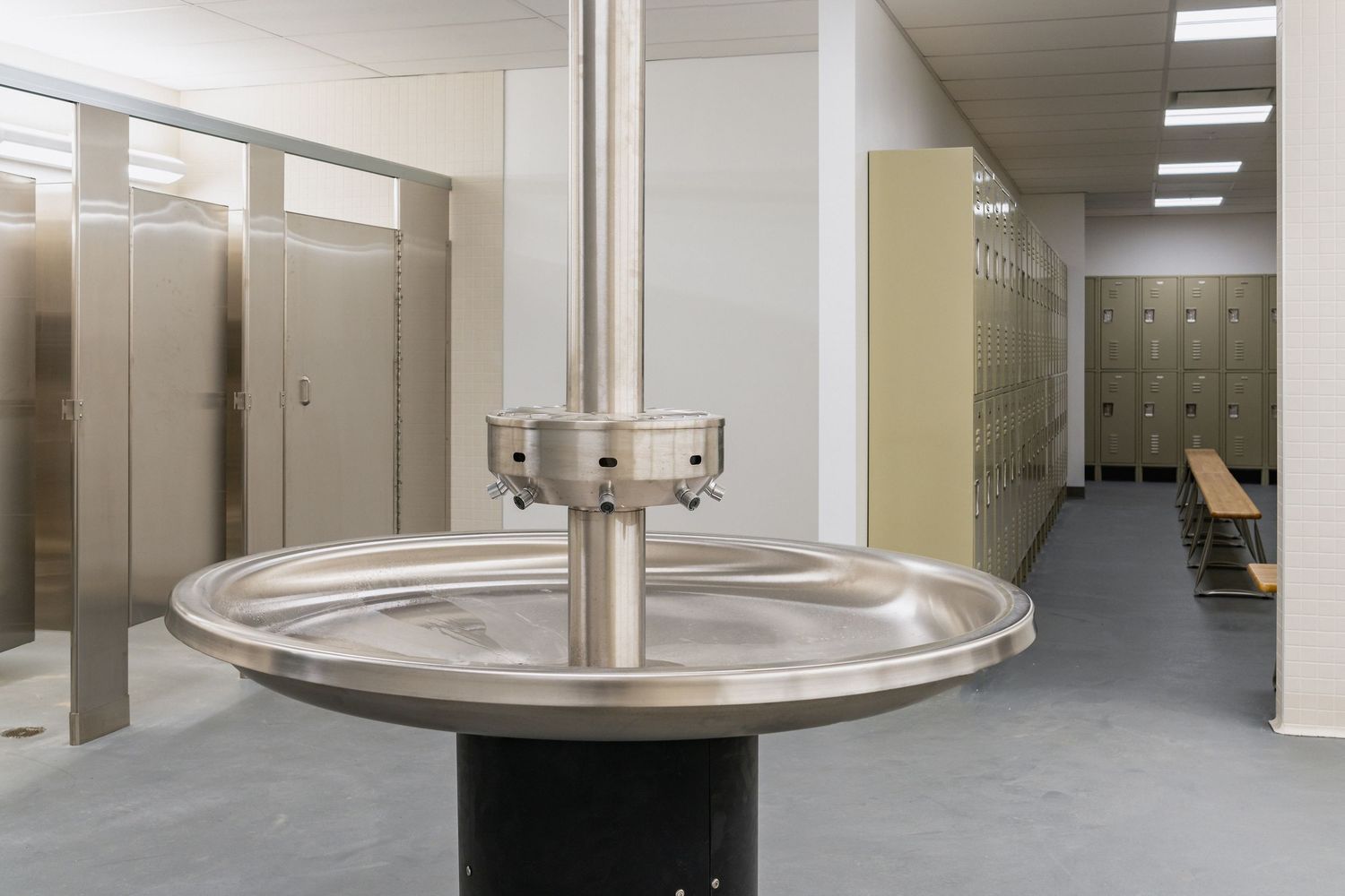 Circular stainless steel wash basin in locker room with bathroom stalls visible in background.