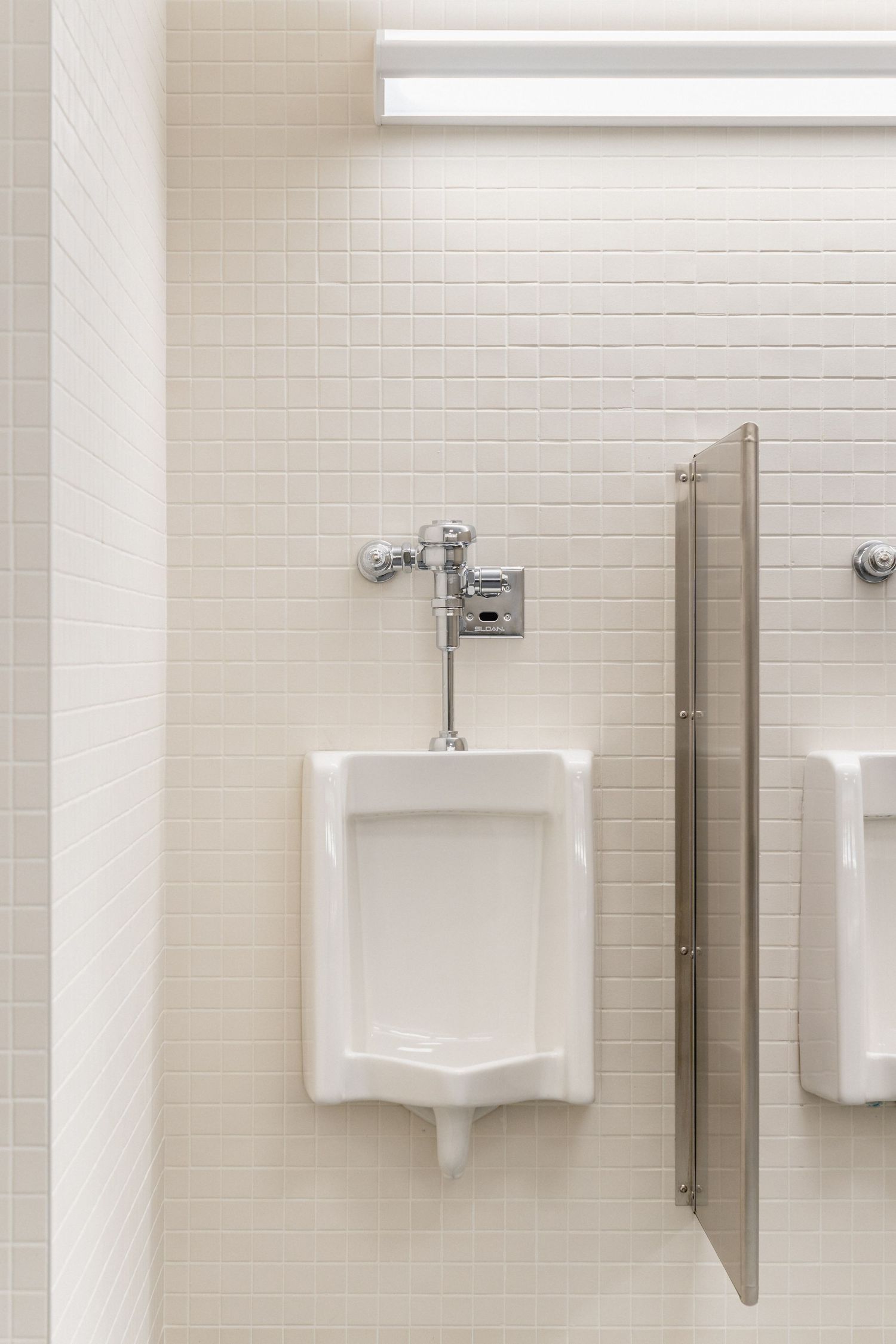 White tiled wall with urinal fixtures and privacy dividers in a commercial bathroom.