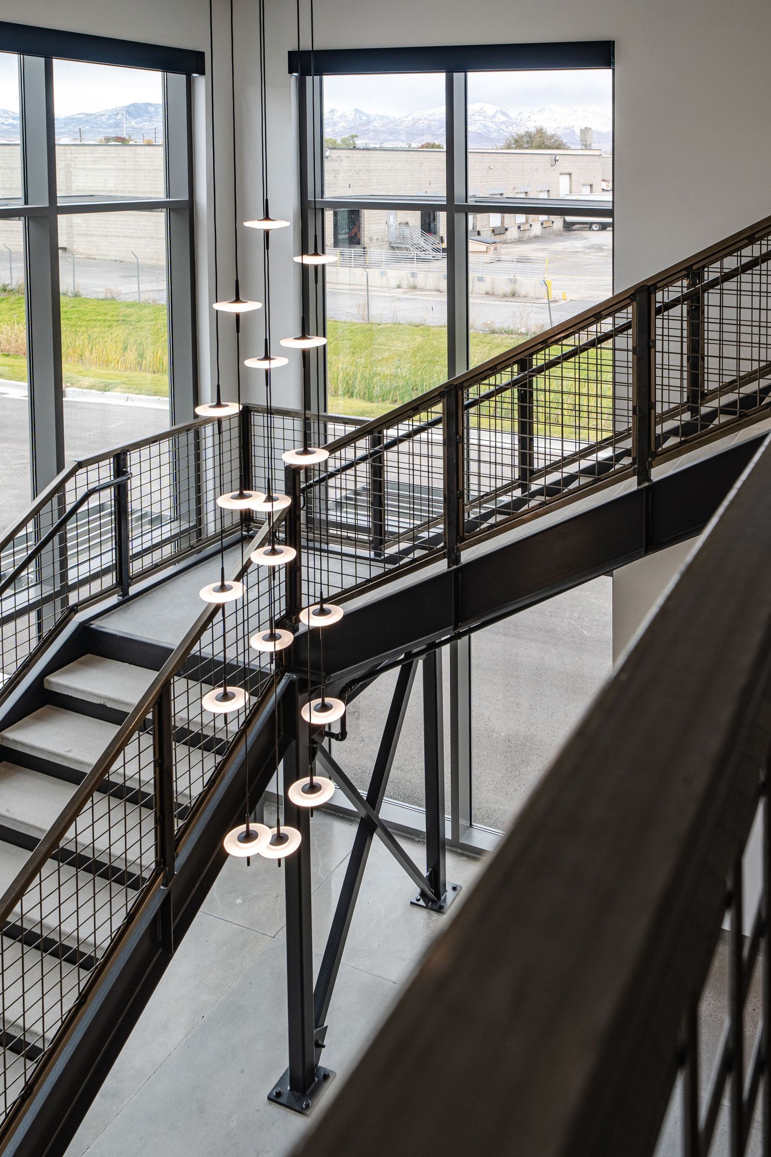 Modern industrial staircase with metal railings and suspended pendant lights near large windows overlooking an urban area.