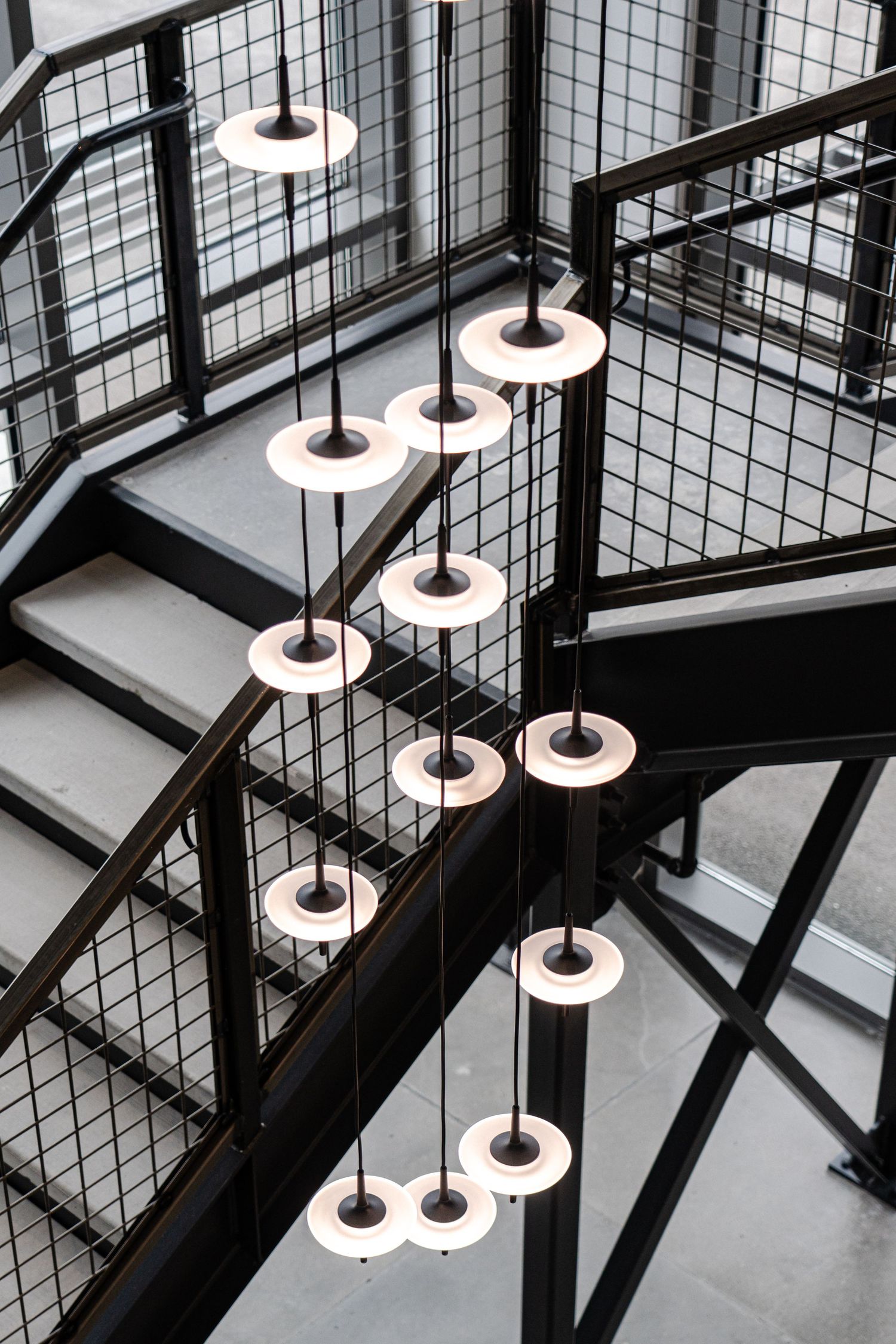 Modern circular pendant lights hang in a cascading pattern along a black industrial staircase with metal mesh railings.