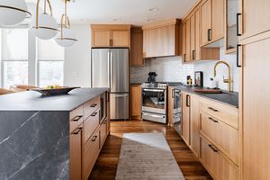 Modern kitchen with light wood cabinets, stainless steel appliances, dark marble countertop, and beige runner rug.