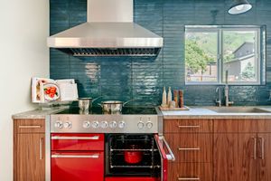 Modern kitchen features teal subway tile backsplash, stainless steel range hood, red stove, and wood cabinets.