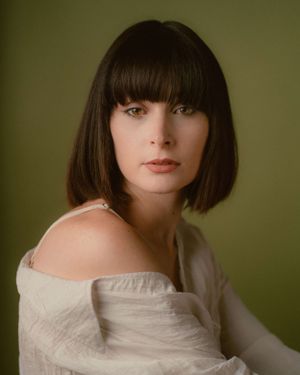 Portrait of a person with dark bangs and white off-shoulder dress against a green background.