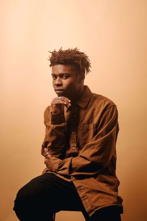 Artistic portrait of a person in a brown jacket against a warm-toned background posing thoughtfully.