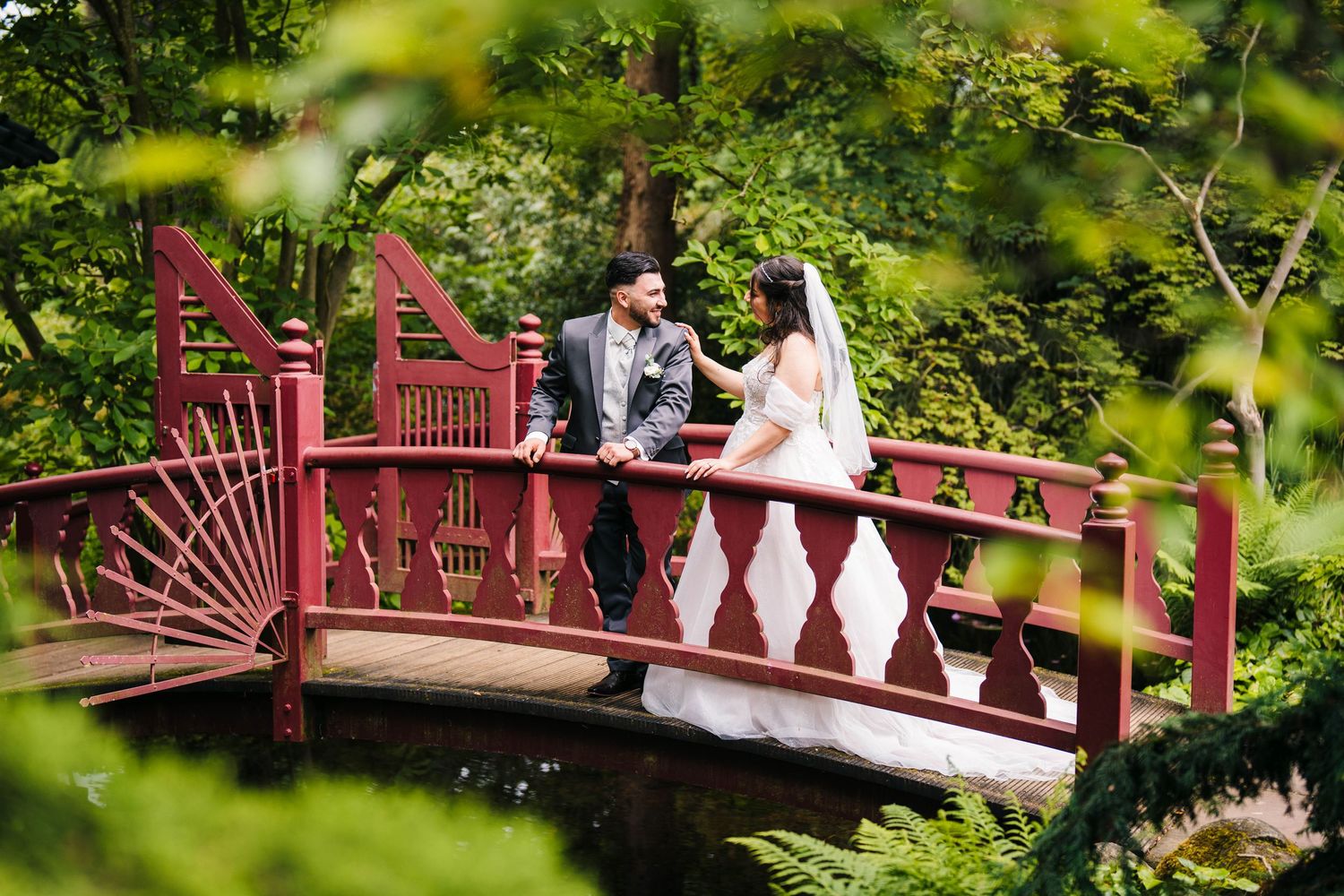Newlyweds share a romantic moment on a red wooden bridge surrounded by lush greenery.