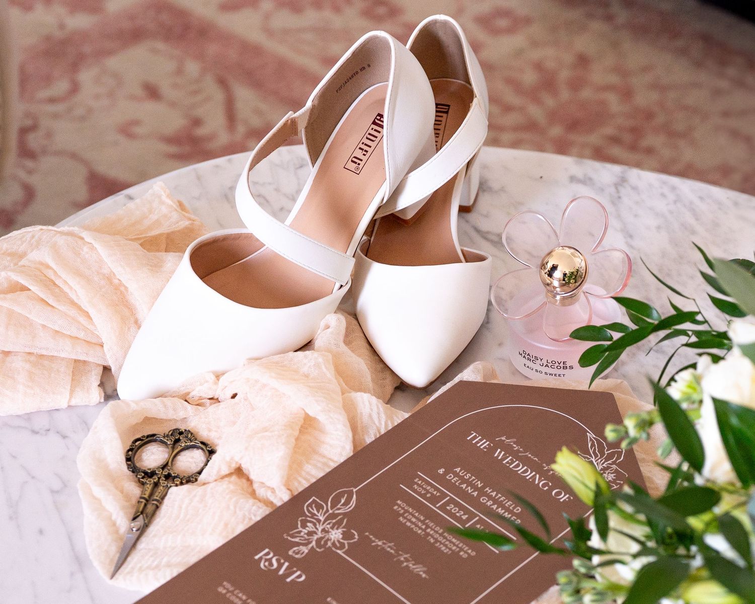 Elegant white bridal shoes with wedding invitation and delicate floral decor on pink fabric.