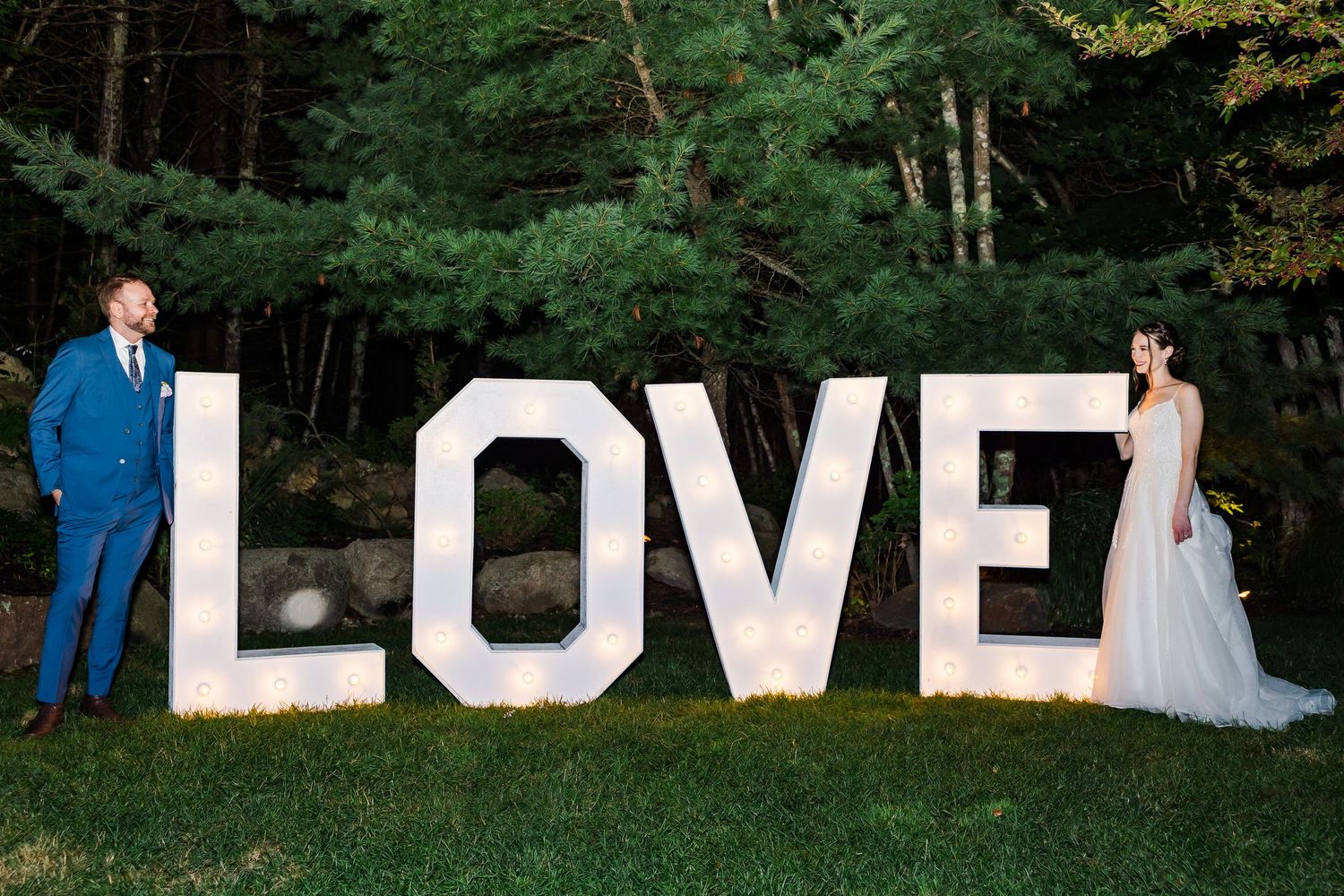 Couple stands with large white LOVE letters display in a garden setting with lush green trees in background.