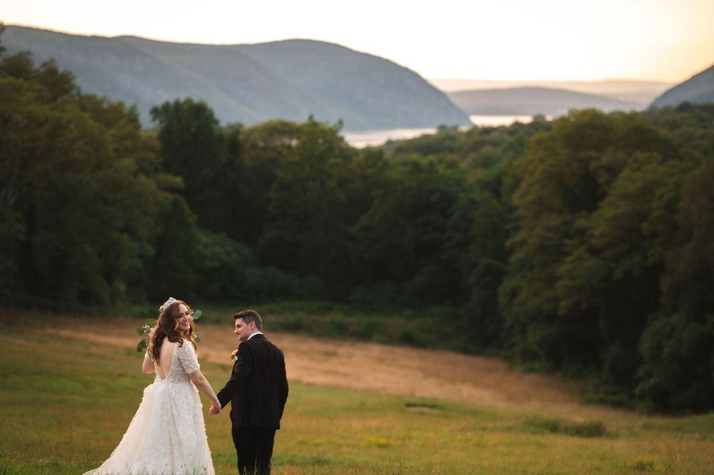 A couple in wedding attire walk through a field at sunset with mountains and the Hudson River in the background of The Garrison in Garrison.