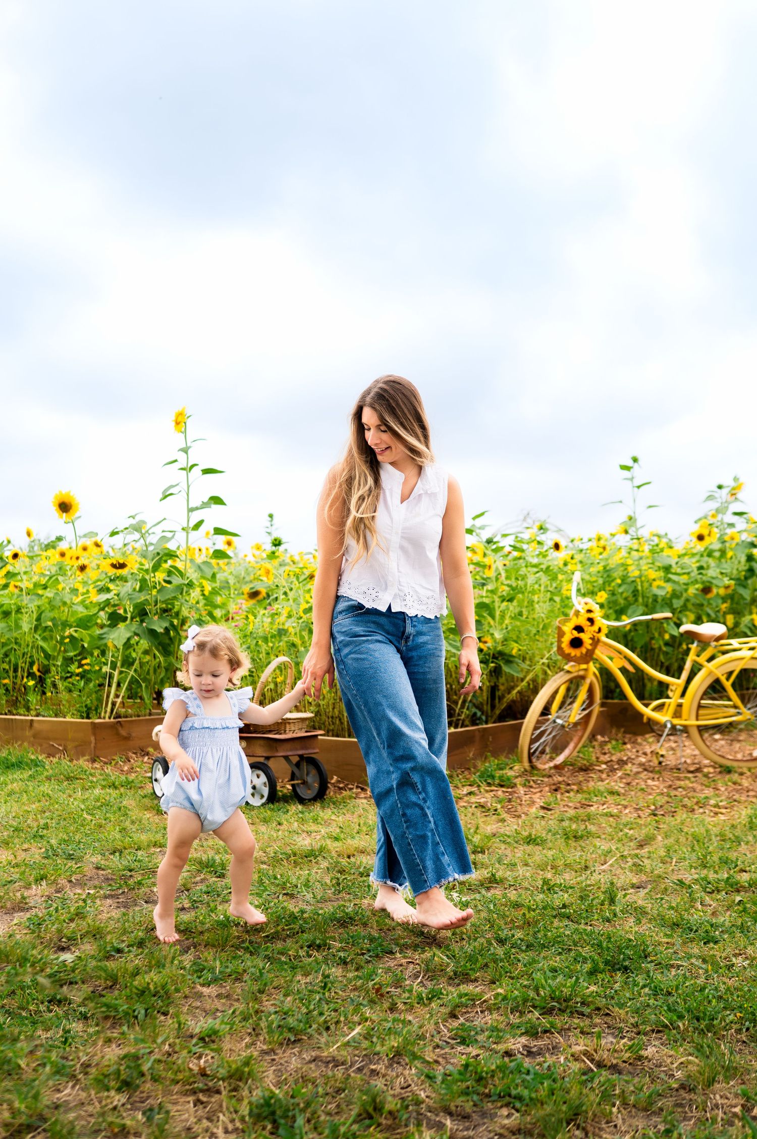 toddler girl holding mothers hand while walking away from sunflower field; matching yellow bicycle in background during family photoshoot