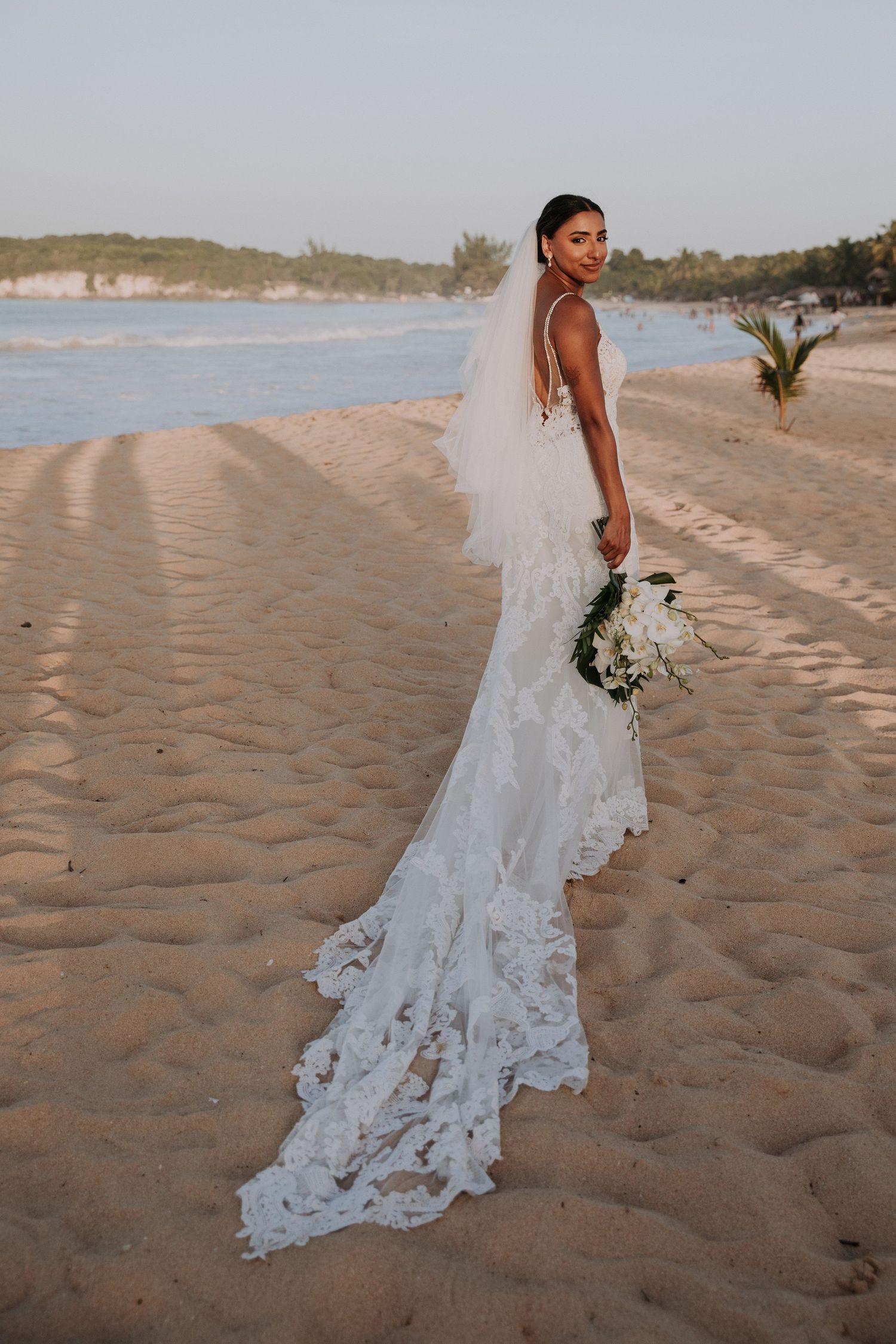 An elegant lace wedding dress train flows across sandy beach during golden hour photoshoot.