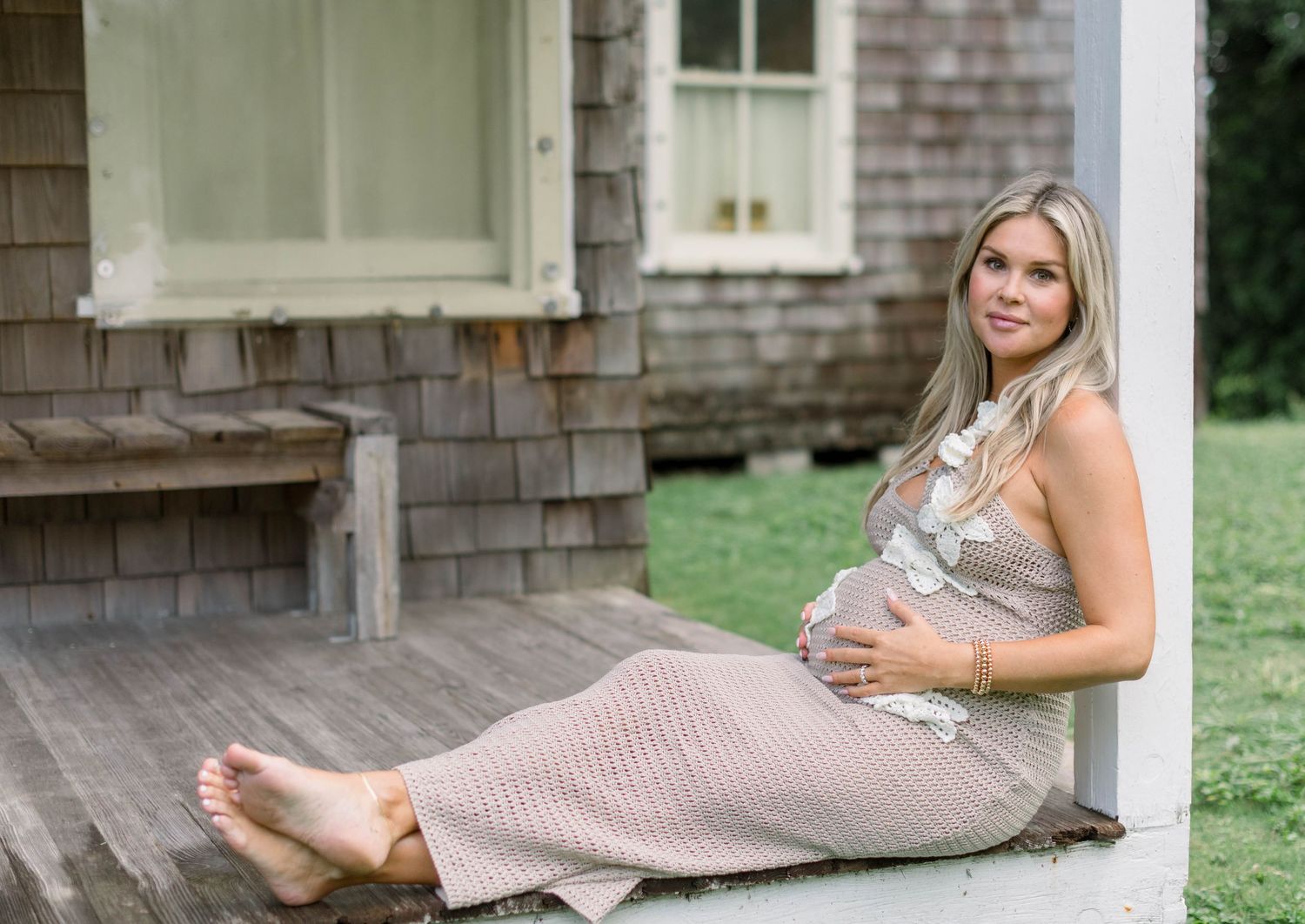 Expectant woman in pink dress reclines on porch steps of rustic shingle-sided house on summer day.