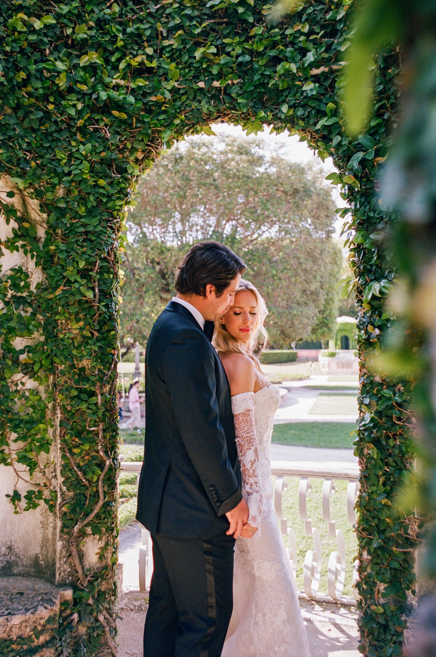 A tender romantic moment shared between a couple among lush ivy-covered columns at a sunlit outdoor venue.