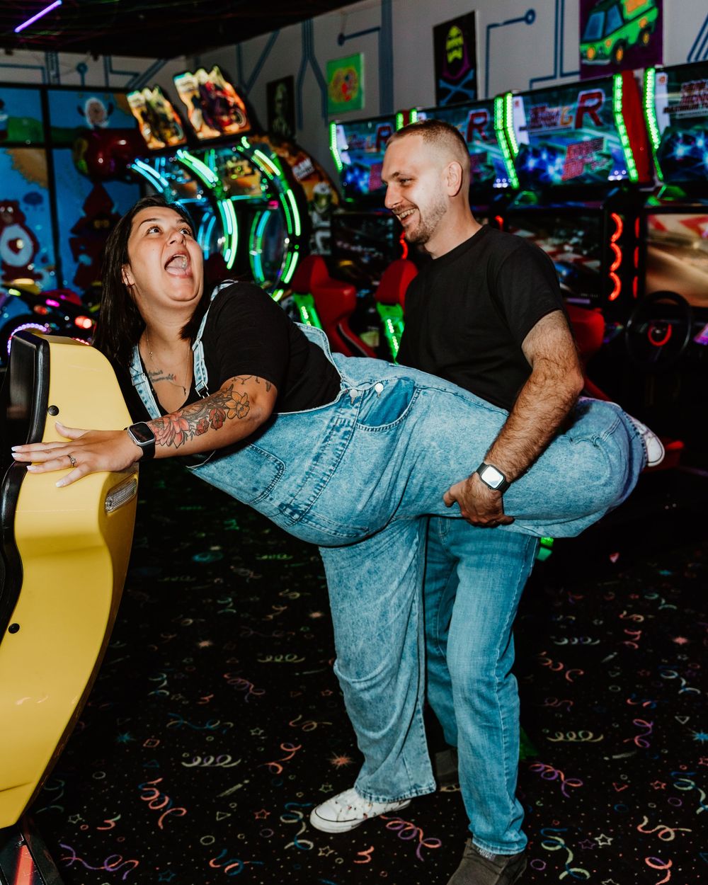 Couple laughing and playing at a colorful arcade, man lifting woman near a yellow racing game machine.