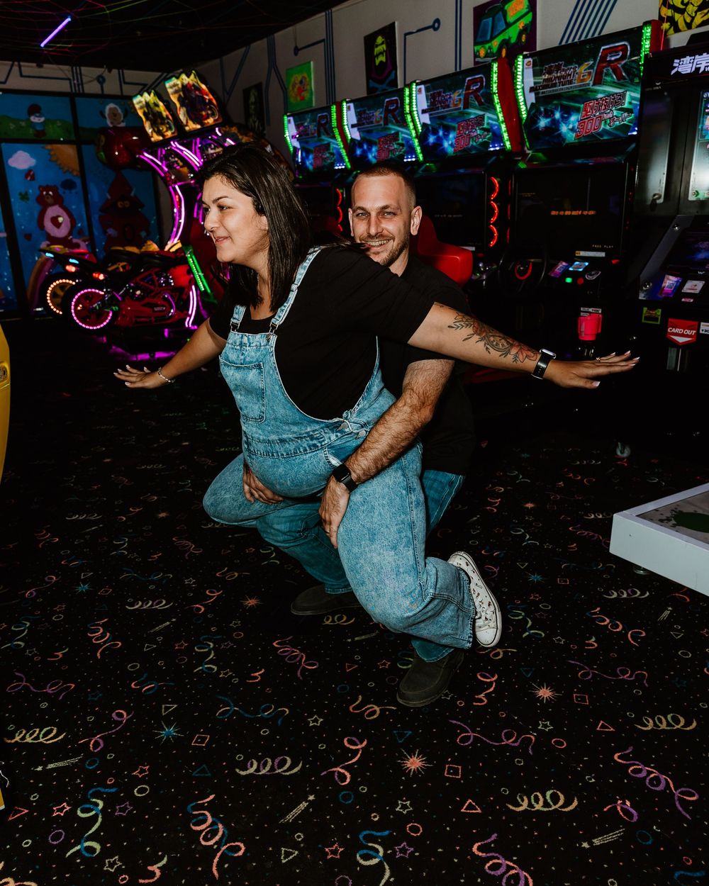 Couple posing playfully in a retro arcade bar, woman in denim overalls and man in black shirt squatting.