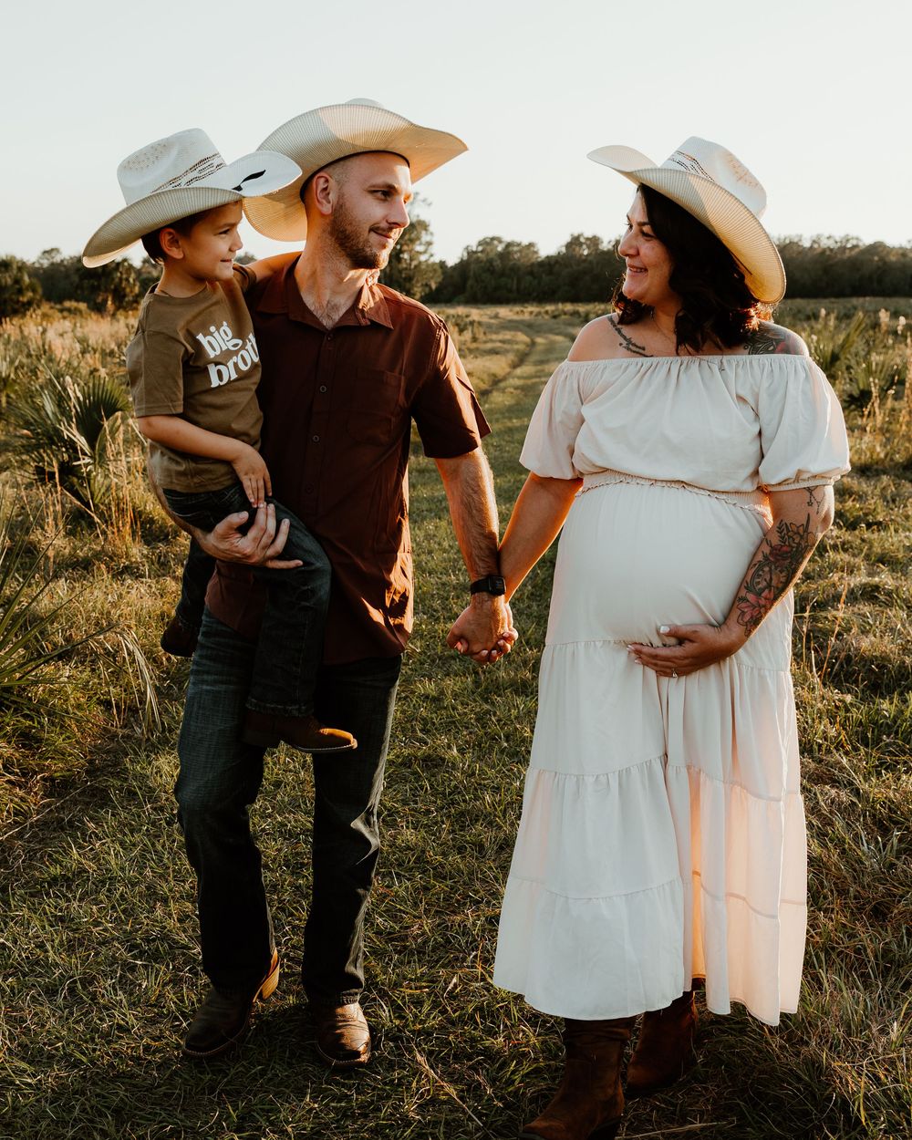 Couple in cowboy hats holding child in sunny field, pregnant woman in white dress cradling baby bump.
