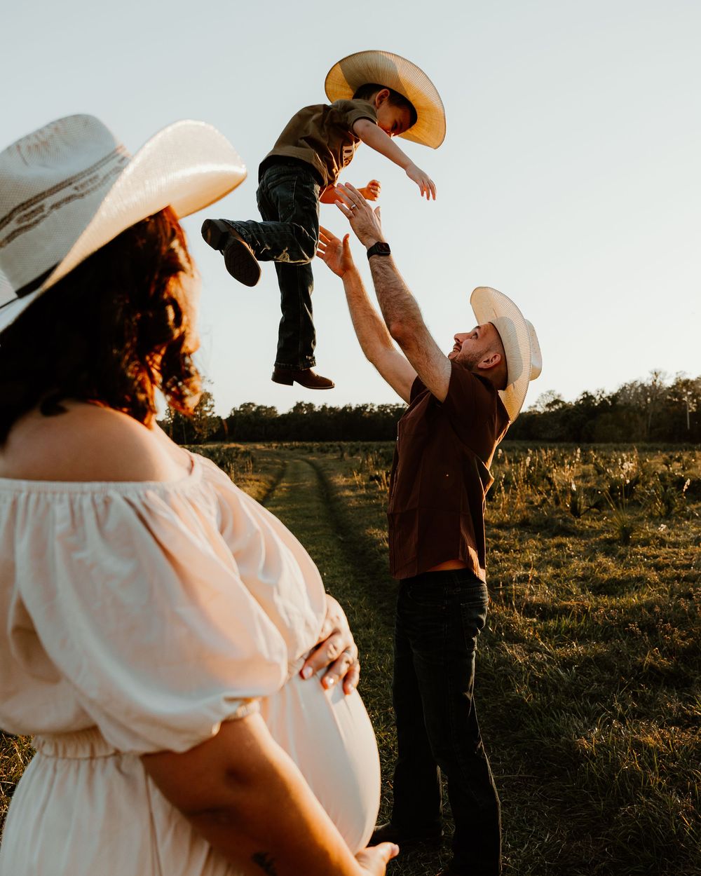 Pregnant woman in white dress and cowboy hat watches man toss young child in air during golden hour family photo.