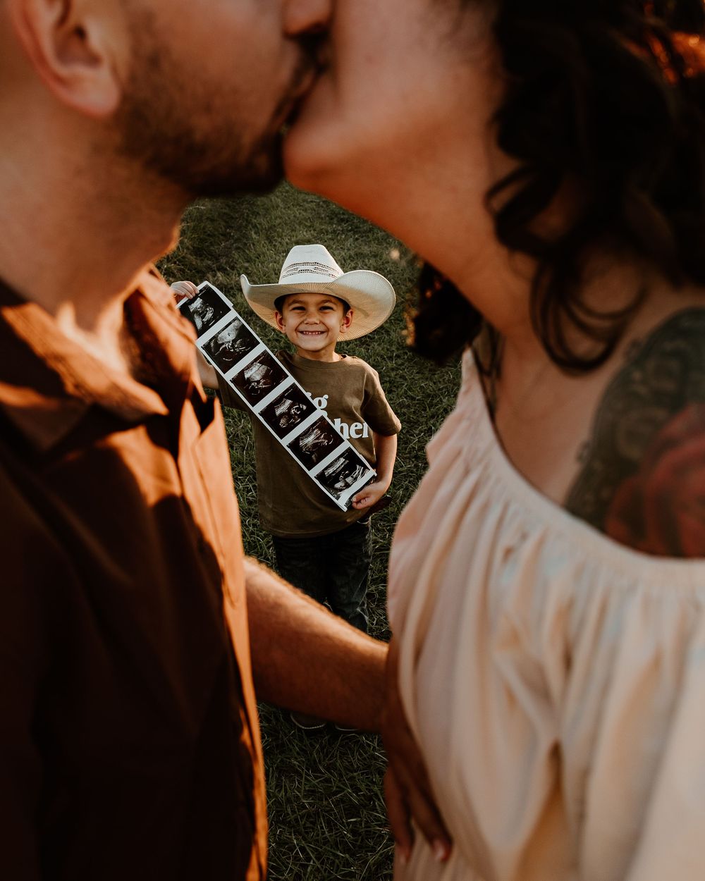 Couple kissing outdoors while smiling child in cowboy hat holds ultrasound photos for pregnancy announcement.